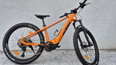 Ein orangefarbenes KTM E-Bike mit schwarzen Rädern und Dämpfern steht vor einer Wand.