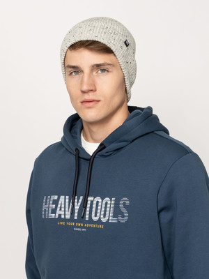 Egy fiatal férfi visel szürke sapkát és kék pulóvert HEAVY TOOLS felirattal.