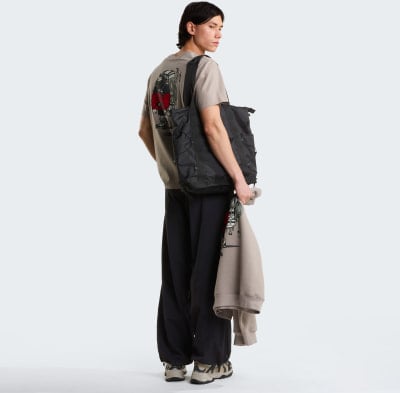 Ein junger Mann in einem beigefarbenen T-Shirt mit Aufdruck und schwarzer Hose trägt eine Tasche.