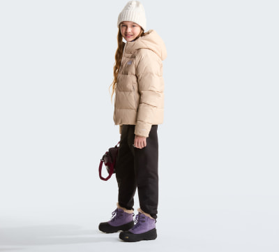 Ein Mädchen in einer beigen Daunenjacke und lila Stiefeln hält eine kleine Handtasche.