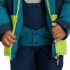 Ein Kind schließt seine Jacke mit einem leuchtend grünen Reißverschluss und dunkelblauen Latzhosen.