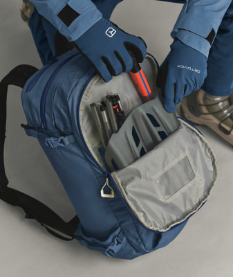 Blaue ORTOVOX Jacke und Handschuhe, blauer Rucksack mit Ausrüstung darin.