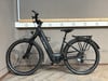 Ein schwarzes E-Bike mit Schutzblechen und Gepäckträger steht auf Holzboden.