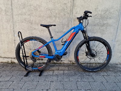 Ein blaues Mountain-E-Bike steht auf einem Ständer vor einer Betonwand. Ein blaues Mountain-E-Bike steht auf einem Ständer vor einer Betonwand.