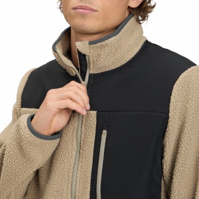 Ein Mann schließt eine beige Fleecejacke mit schwarzen Einsätzen und grauem Besatz. Ein Mann schließt eine beige Fleecejacke mit schwarzen Einsätzen und grauem Besatz.