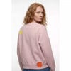 Eine rothaarige Frau trägt einen rosafarbenen Pullover mit gelbem und orangem Aufnäher.