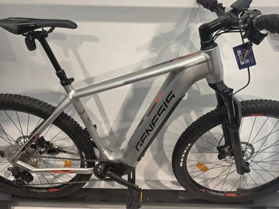 Ein silbernes E-Bike von Genesis mit orangefarbenen Akzenten steht auf einem Ständer. Ein silbernes E-Bike von Genesis mit orangefarbenen Akzenten steht auf einem Ständer.