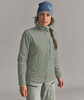 Eine Frau in einer blauen Mütze und einer grünen Jacke steht vor einem grauen Hintergrund.