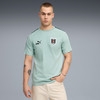 Ein junger Mann in einem hellblauen T-Shirt mit Logo und Schriftzug posiert vor grauem Hintergrund.