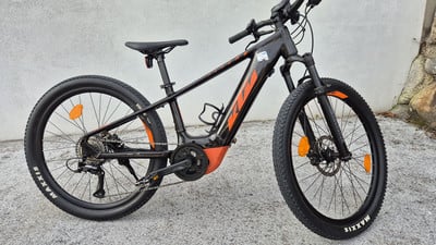Ein schwarzes KTM E-Bike mit orangefarbenen Details steht vor einer Steinmauer. Ein schwarzes KTM E-Bike mit orangefarbenen Details steht vor einer Steinmauer.