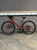 Ein orangefarbenes Gravelbike steht auf einem Holzboden vor einer grauen Wand.