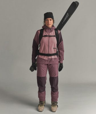Eine Frau in Skikleidung mit Skiern auf dem Rücken vor grauem Hintergrund.