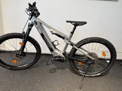 Ein silbernes E-Mountainbike mit Federung und Scheibenbremsen auf dunklem Teppich. Ein silbernes E-Mountainbike mit Federung und Scheibenbremsen auf dunklem Teppich.