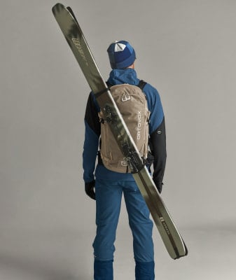 Ein Mann mit Skiern auf dem Rücken und blauer Hose steht vor grauem Hintergrund. Ein Mann mit Skiern auf dem Rücken und blauer Hose steht vor grauem Hintergrund.