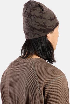 Ein Mann mit langen dunklen Haaren trägt eine braune Strickmütze mit Camouflage-Muster.