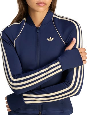 Dunkelblaue Adidas Jacke für Damen mit cremefarbenen Streifen und Logo auf der Brust. Dunkelblaue Adidas Jacke für Damen mit cremefarbenen Streifen und Logo auf der Brust.