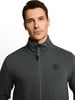 Egy férfi sötétzöld, álló galléros, cipzáros dzsekiben.