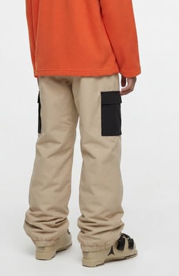Mann in einem orangefarbenen Pullover und beigen Cargohosen mit schwarzen Taschen und Skischuhen.