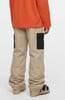 Mann in einem orangefarbenen Pullover und beigen Cargohosen mit schwarzen Taschen und Skischuhen.