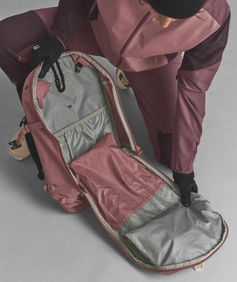 Eine Person in einer rosafarbenen Skihose öffnet einen Rucksack mit vielen Taschen.