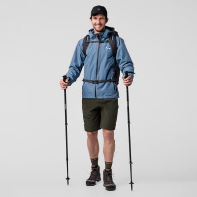 Ein Mann in blauer Jacke und Shorts hält Wanderstöcke.