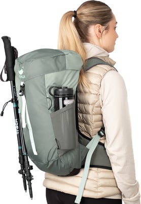 Eine Frau mit einem Rucksack und Wanderstöcken, die eine beige Weste trägt.