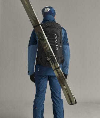Ein Skifahrer mit Rucksack und Skiern auf dem Rücken vor grauem Hintergrund. Ein Skifahrer mit Rucksack und Skiern auf dem Rücken vor grauem Hintergrund.