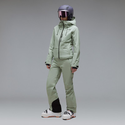 Eine Skifahrerin in grüner Kleidung und Helm mit Brille vor grauem Hintergrund.
