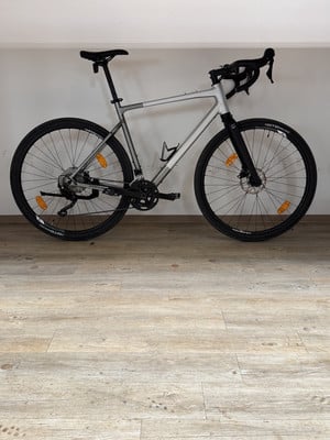 Ein silbernes Gravelbike mit schwarzem Lenker und Reifen steht auf einem Holzboden. Ein silbernes Gravelbike mit schwarzem Lenker und Reifen steht auf einem Holzboden.