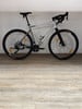 Ein silbernes Gravelbike mit schwarzem Lenker und Reifen steht auf einem Holzboden.