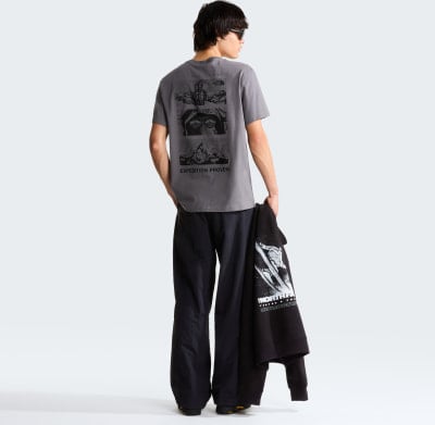 Ein Mann in einem grauen T-Shirt mit Bergmotiv und schwarzer Hose hält einen Hoodie. Ein Mann in einem grauen T-Shirt mit Bergmotiv und schwarzer Hose hält einen Hoodie.