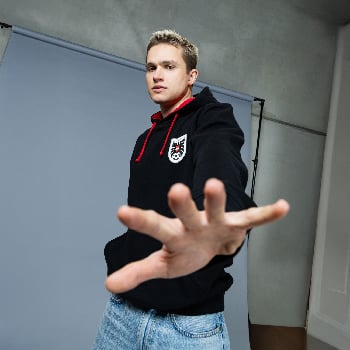 Ein junger Mann im schwarzen Hoodie und blauer Jeans blickt direkt in die Kamera.