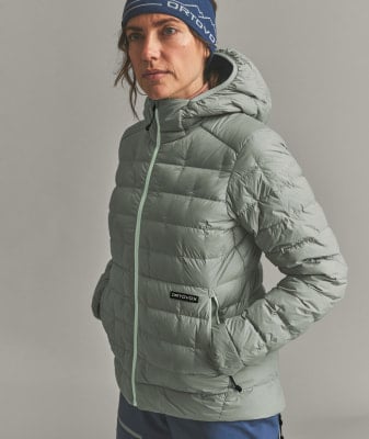 Eine Frau in einer Daunenjacke und Mütze mit ORTOVOX-Logo vor grauem Hintergrund. Eine Frau in einer Daunenjacke und Mütze mit ORTOVOX-Logo vor grauem Hintergrund.