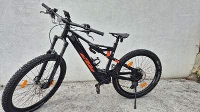 Ein schwarzes KTM E-Mountainbike mit orangefarbenen Akzenten steht vor einer Wand. Ein schwarzes KTM E-Mountainbike mit orangefarbenen Akzenten steht vor einer Wand.