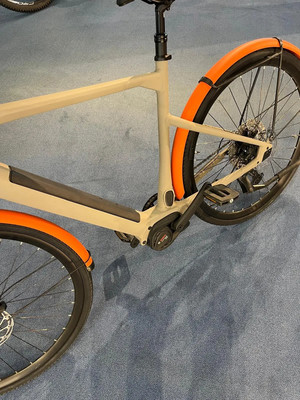 Ein beiges E-Bike mit orangen Schutzblechen auf blauem Teppich.