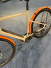 Ein beiges E-Bike mit orangen Schutzblechen auf blauem Teppich.