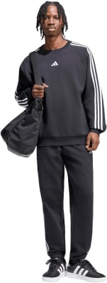 Ein Mann in schwarzer Adidas-Kleidung mit weißen Streifen und einer Umhängetasche.