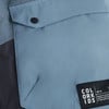 Detail einer Taschenklappe und eines Logos auf einer blauen Jacke.