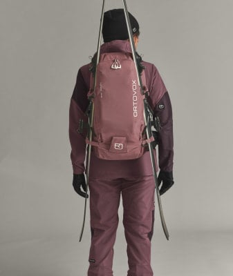Rückansicht einer Person mit Rucksack und Skiern auf grauem Hintergrund.