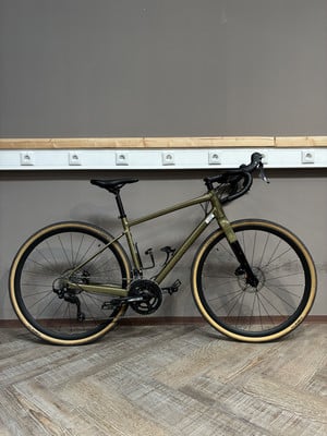 Ein grünes Gravelbike steht auf einem Holzboden vor einer grauen Wand mit Steckdosen. Ein grünes Gravelbike steht auf einem Holzboden vor einer grauen Wand mit Steckdosen.