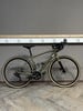 Ein grünes Gravelbike steht auf einem Holzboden vor einer grauen Wand mit Steckdosen.