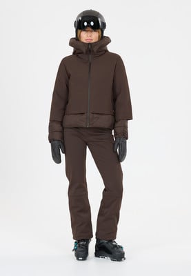 Eine Frau in brauner Skijacke, Hose, Helm und Handschuhen steht vor weißem Hintergrund. Eine Frau in brauner Skijacke, Hose, Helm und Handschuhen steht vor weißem Hintergrund.