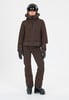 Eine Frau in brauner Skijacke, Hose, Helm und Handschuhen steht vor weißem Hintergrund.