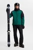 Ein Skifahrer hält Skier, trägt eine grüne Jacke und schwarze Hose.