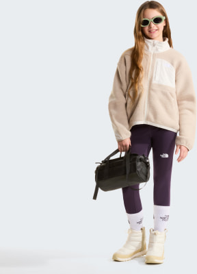 Ein Mädchen in einer beige Fleecejacke und lila Leggings hält eine schwarze Tasche.