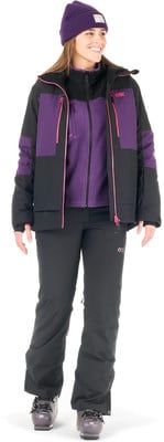 Eine Frau in einer Skijacke und Hose mit einer lila Mütze und einem Fleece-Pullover.