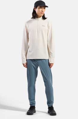 Ein Mann mit schwarzer Kappe trägt einen beigen Pullover und blaue Hose.