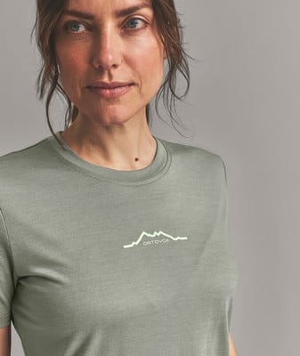 Eine Frau in einem grünen T-Shirt mit einem Berglogo und dem Schriftzug ORTOVOX. Eine Frau in einem grünen T-Shirt mit einem Berglogo und dem Schriftzug ORTOVOX.