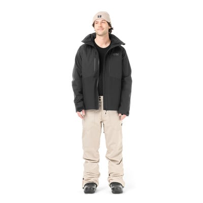 Ein Mann in einer schwarzen Jacke, beige Hose und Mütze steht vor weißem Hintergrund.