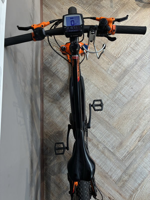 Ein schwarzes E-Bike mit orangefarbenen Details und Digitalanzeige steht an einer Wand.